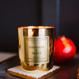 Gold candle with 'Goodness & Mercy' label on a dark surface with a red fruit in the background