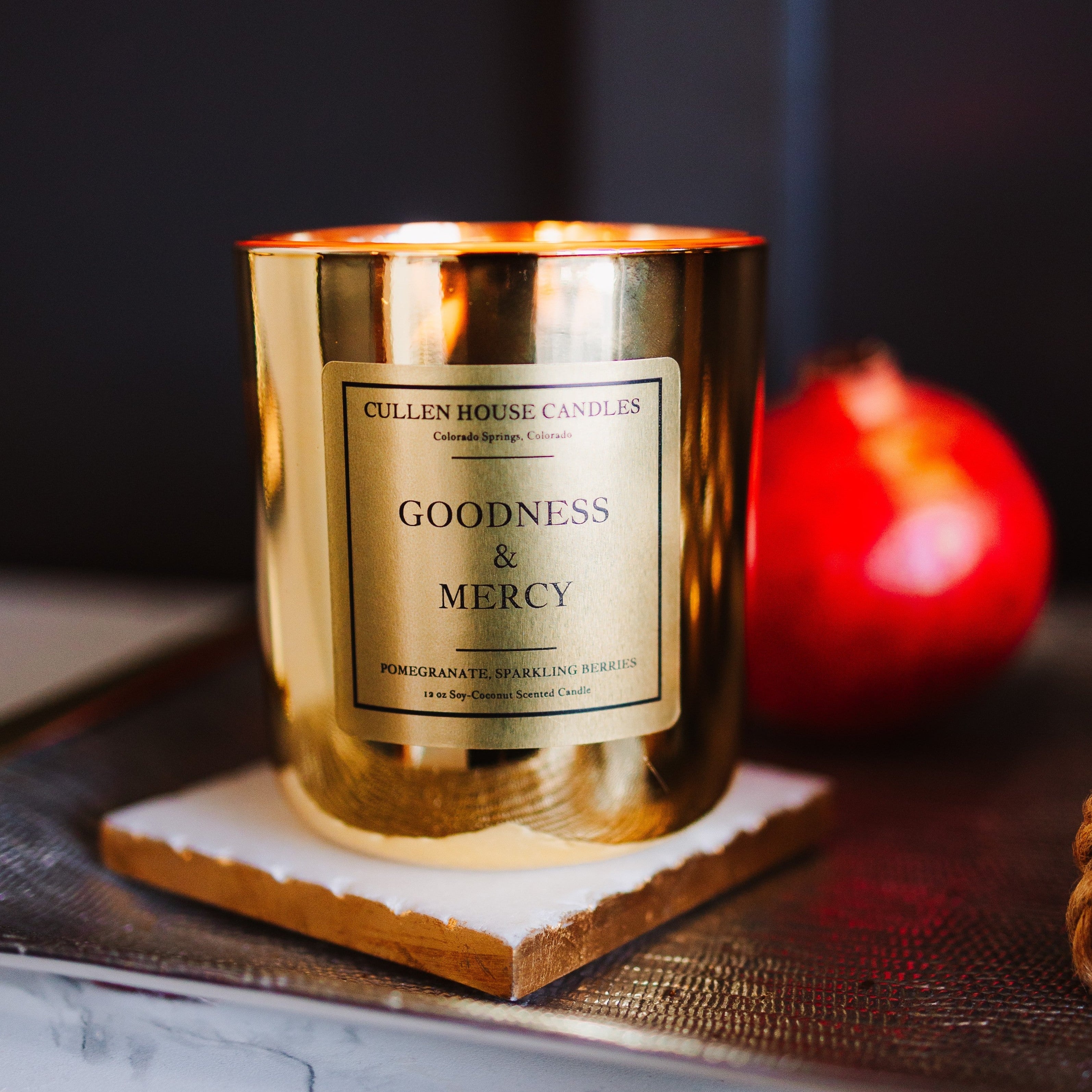 Gold candle with 'Goodness & Mercy' label on a dark surface with a red fruit in the background