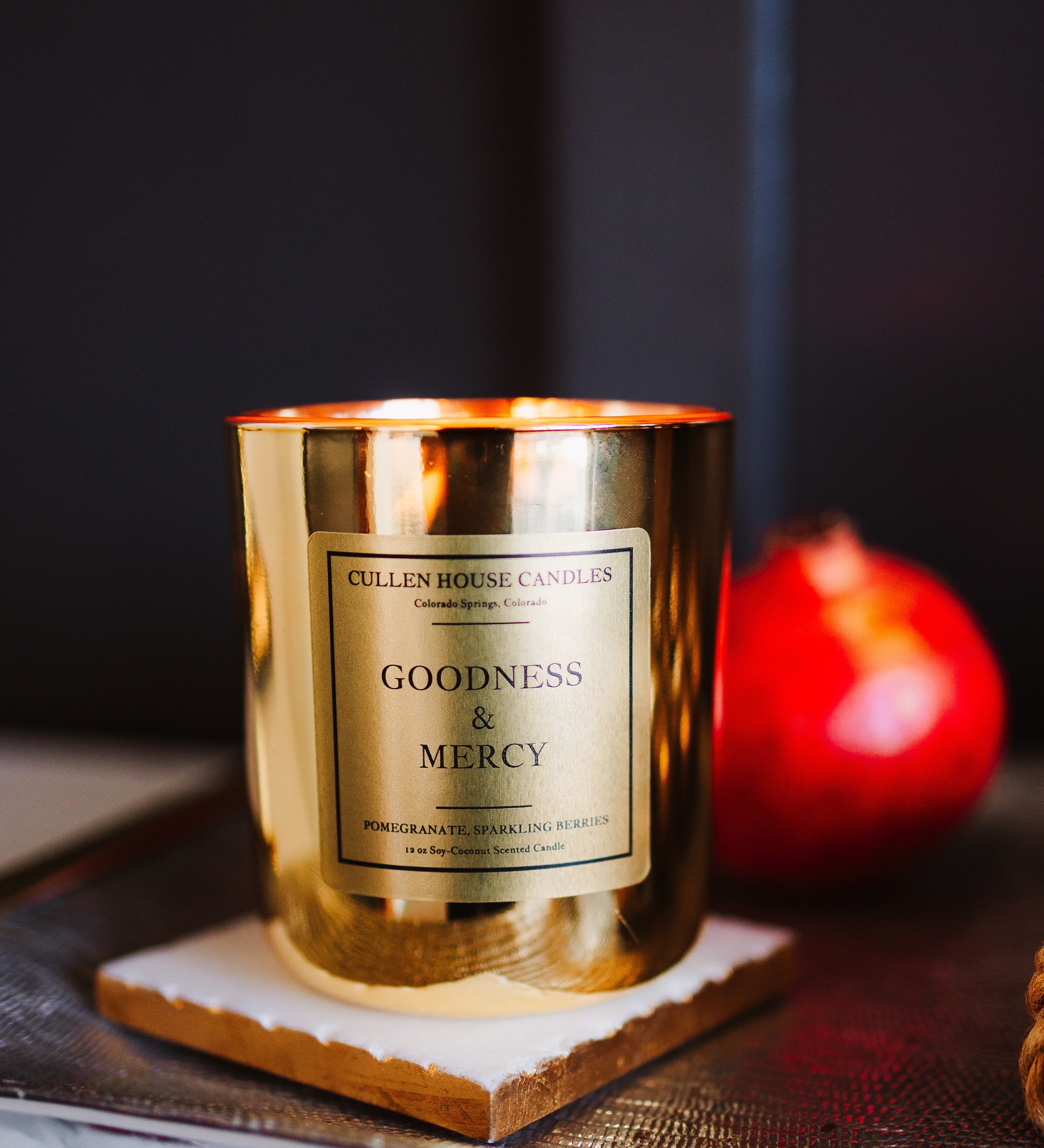 Gold candle with 'Goodness & Mercy' label on a dark surface with a red fruit in the background