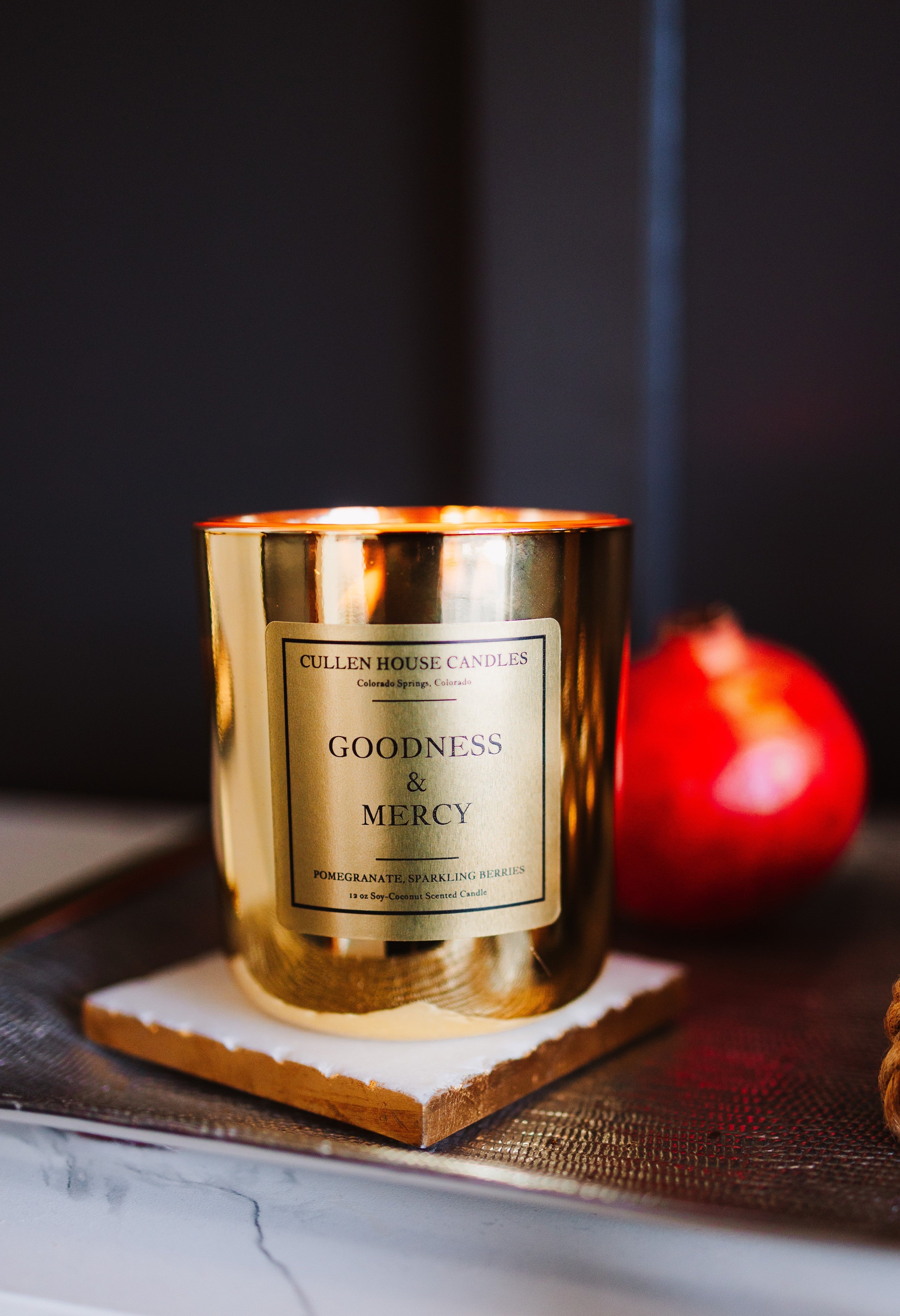 Gold candle with 'Goodness & Mercy' label on a dark surface with a red fruit in the background