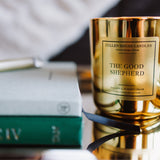 Gold candle labeled 'The Good Shepherd' on a reflective surface with a blurred background