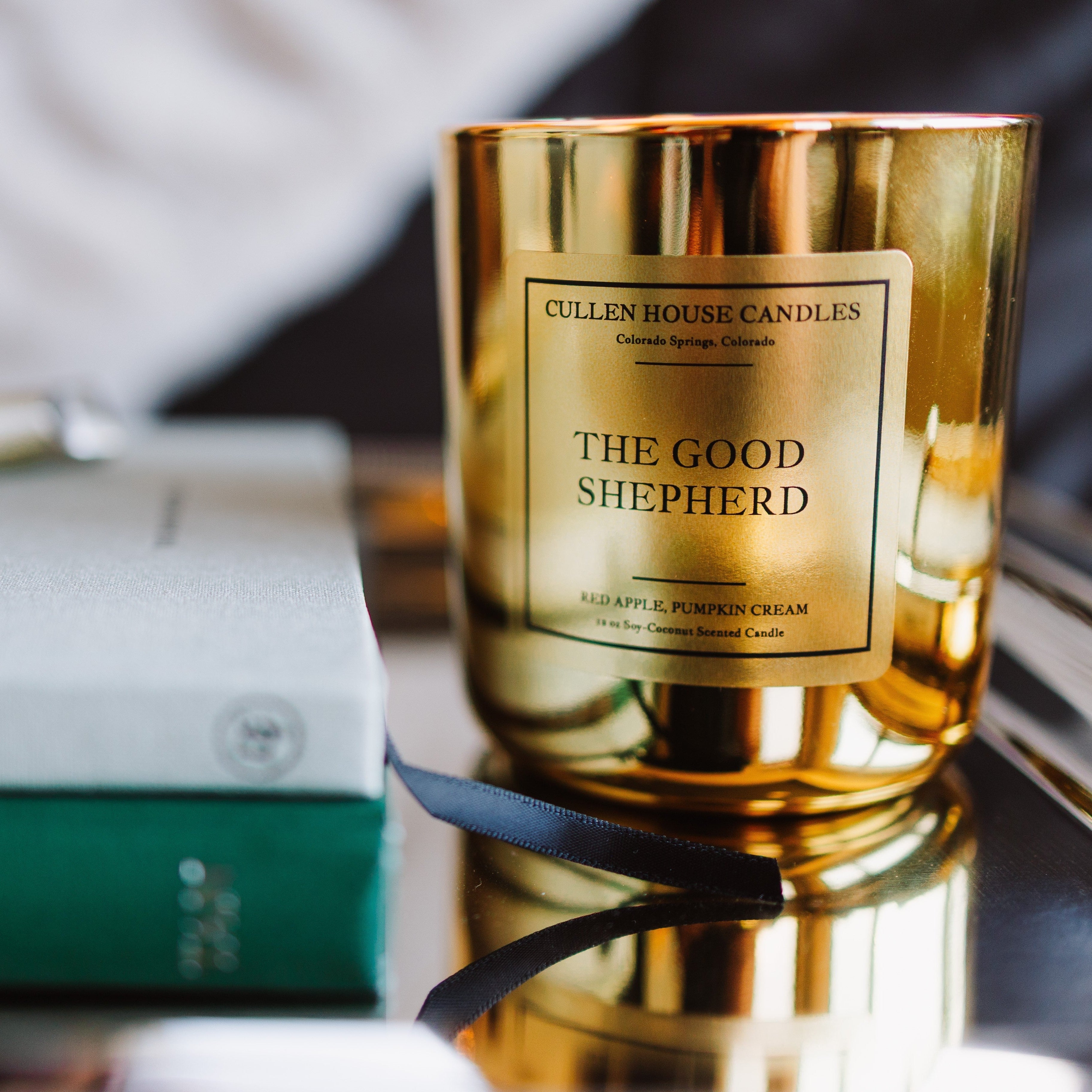 Gold candle labeled 'The Good Shepherd' on a reflective surface with a blurred background