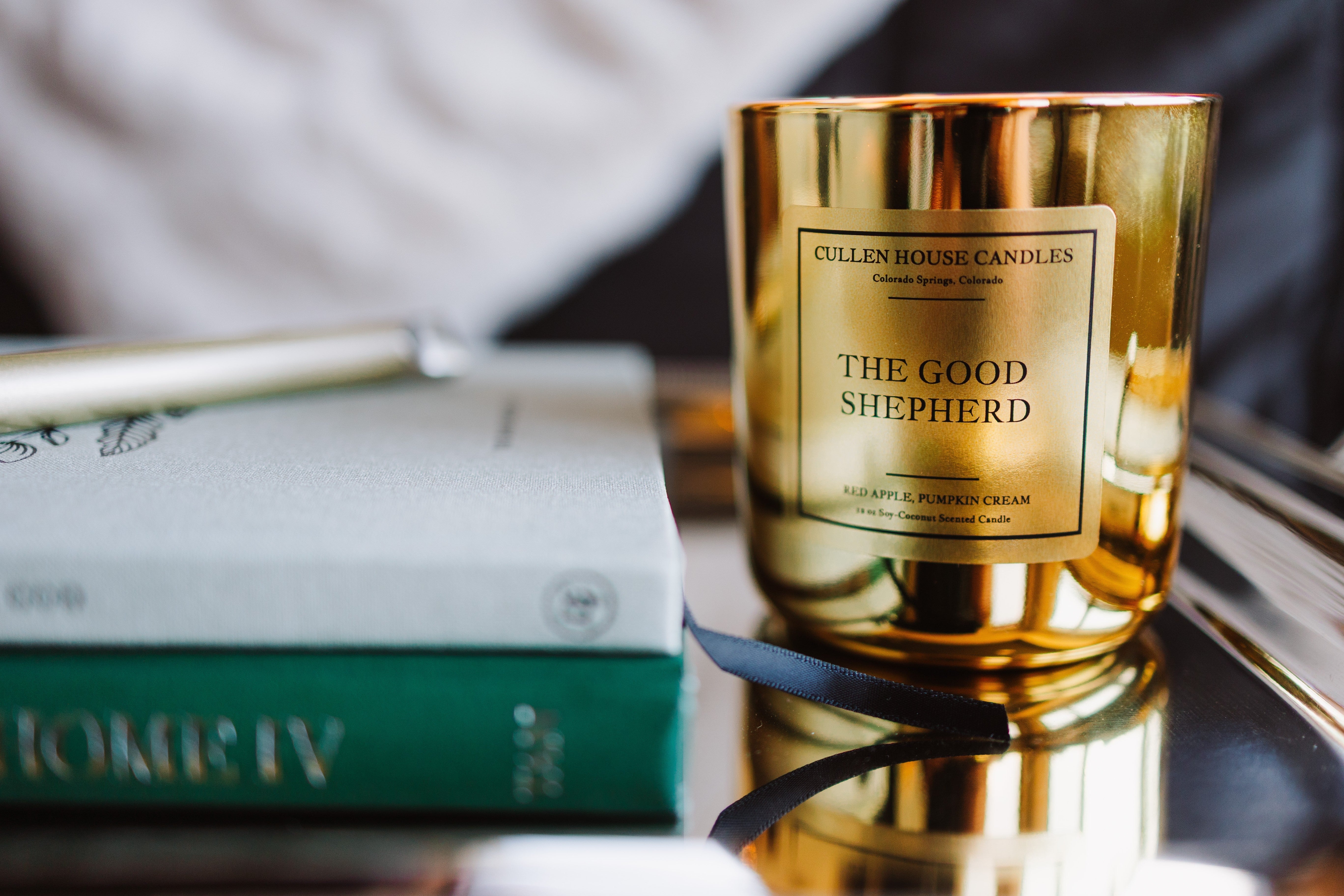 Gold candle labeled 'The Good Shepherd' on a reflective surface with a blurred background