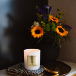 Candle with a decorative label on a tray next to a vase of flowers on a marble surface.