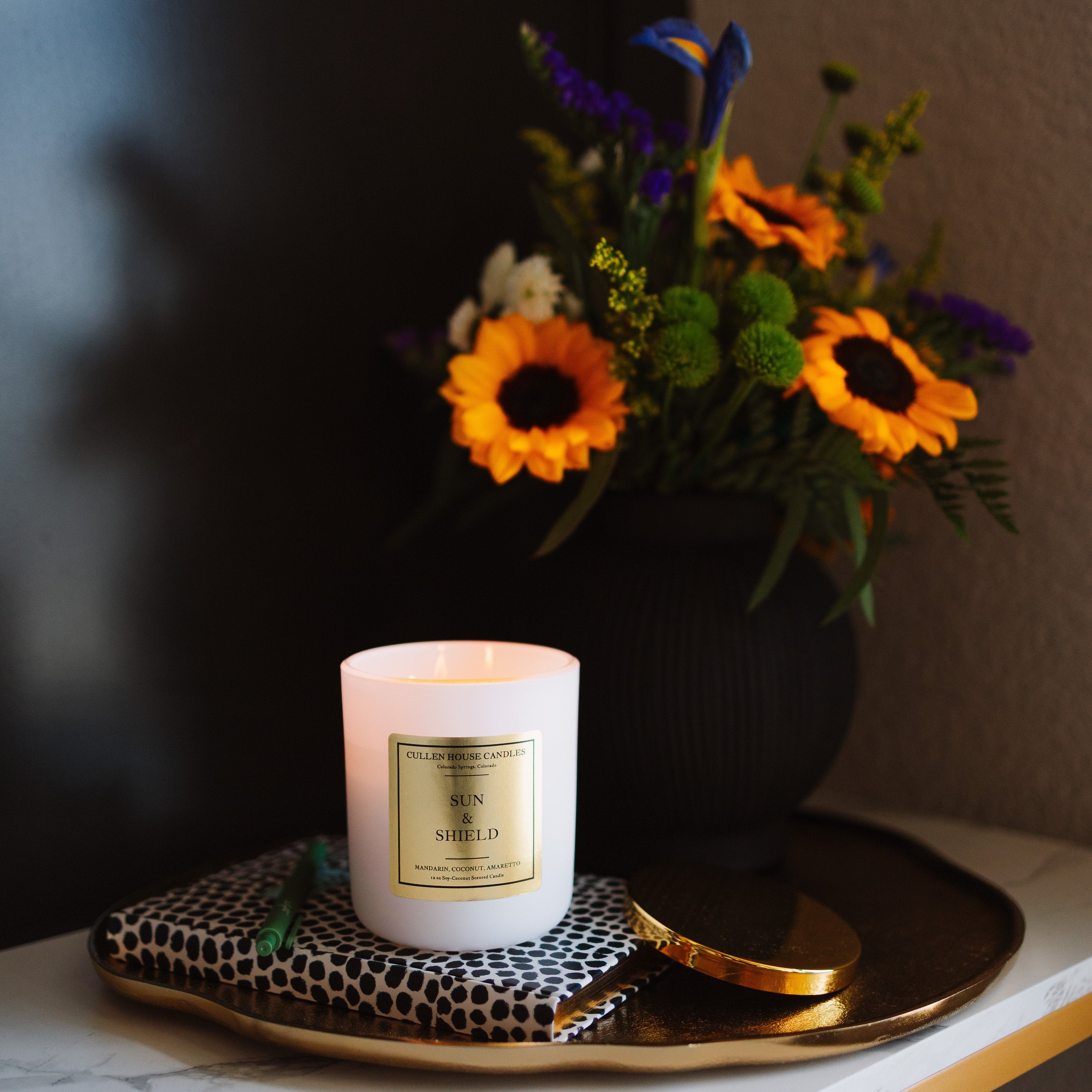 Candle with a decorative label on a tray next to a vase of flowers on a marble surface.