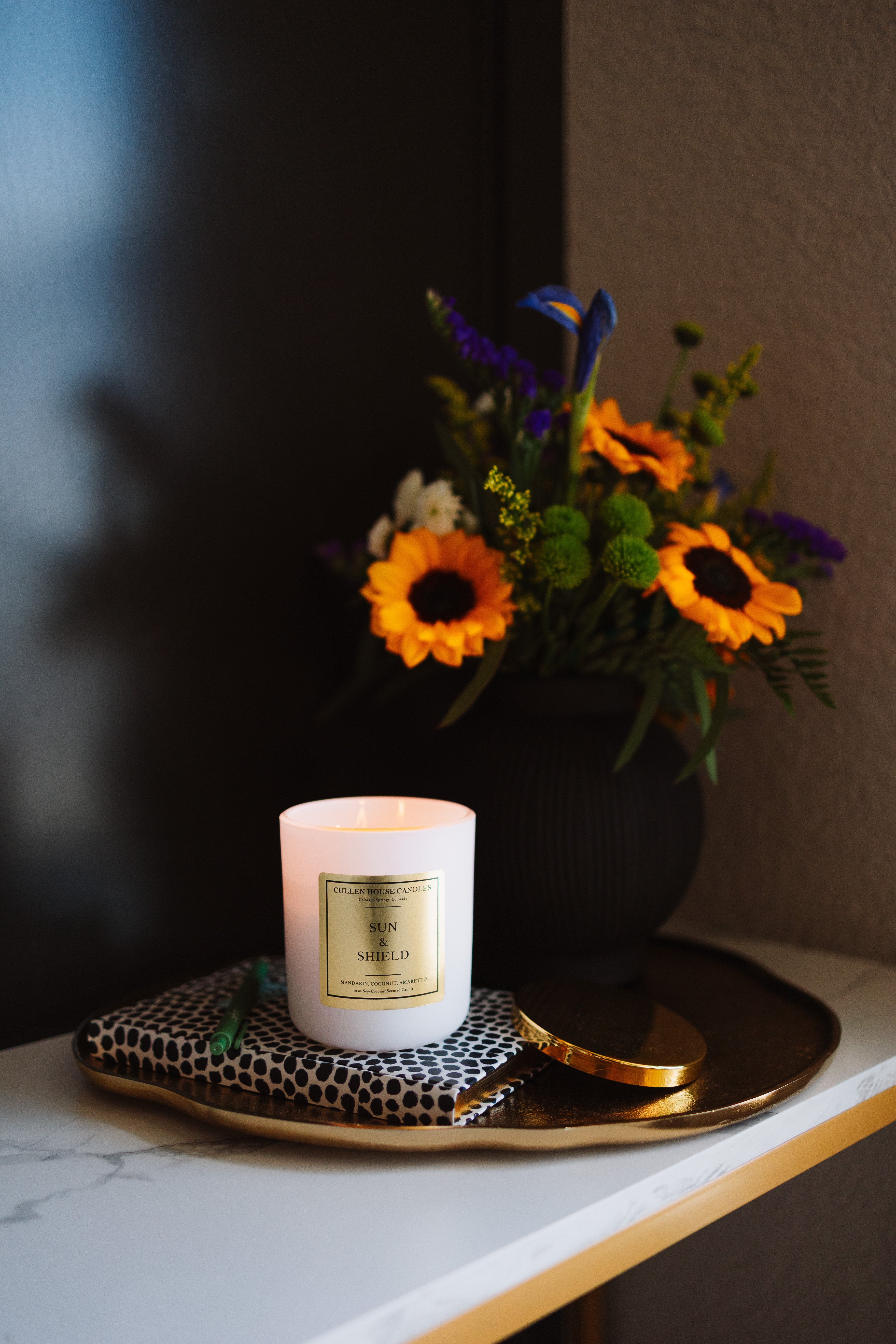 Candle with a decorative label on a tray next to a vase of flowers on a marble surface.