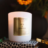 Candle labeled 'Sun & Shield' on a decorative surface with flowers in the background