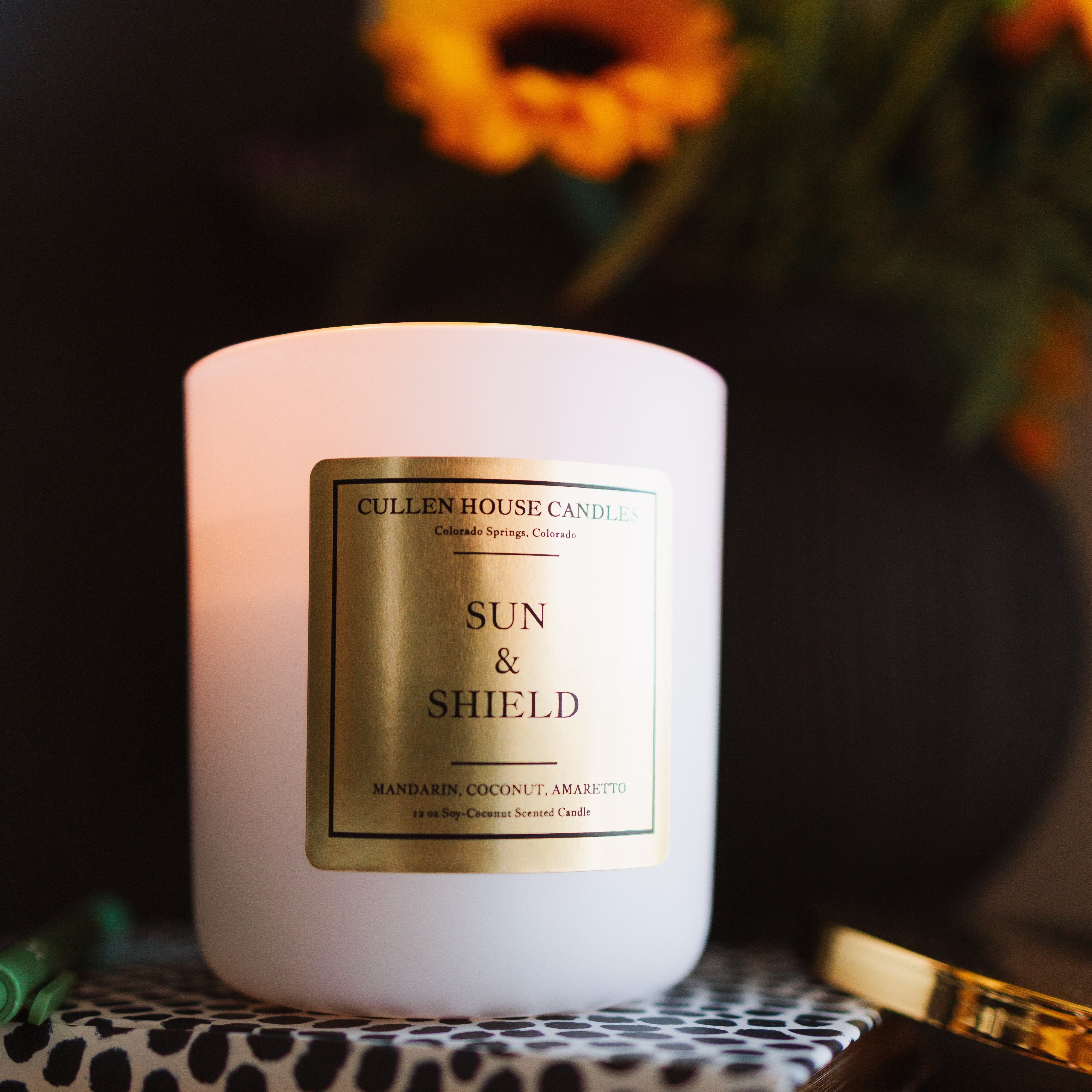 Candle labeled 'Sun & Shield' on a decorative surface with flowers in the background