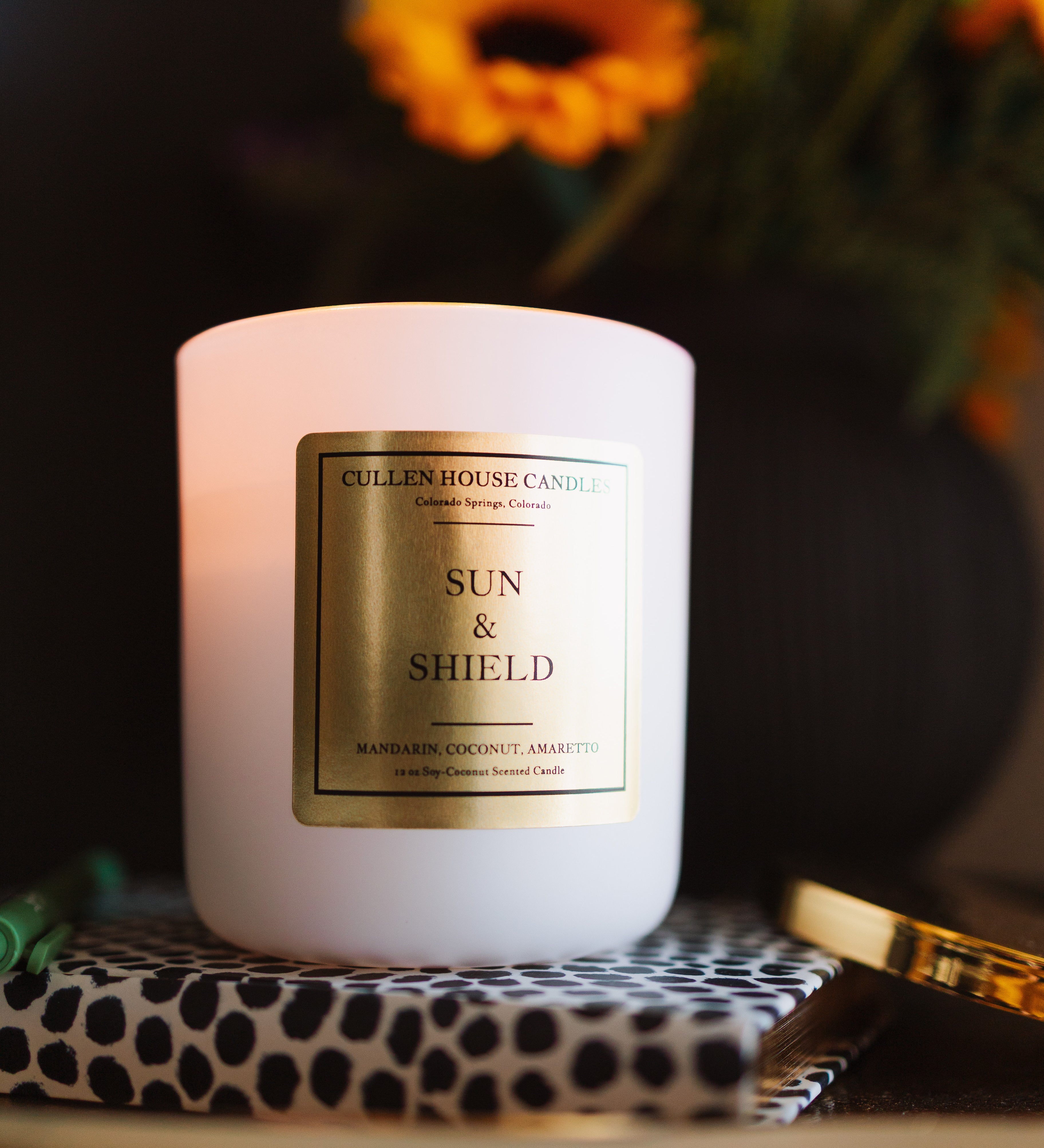 Candle labeled 'Sun & Shield' on a decorative surface with flowers in the background