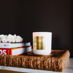 White candle with gold label on a woven tray with books against a dark background