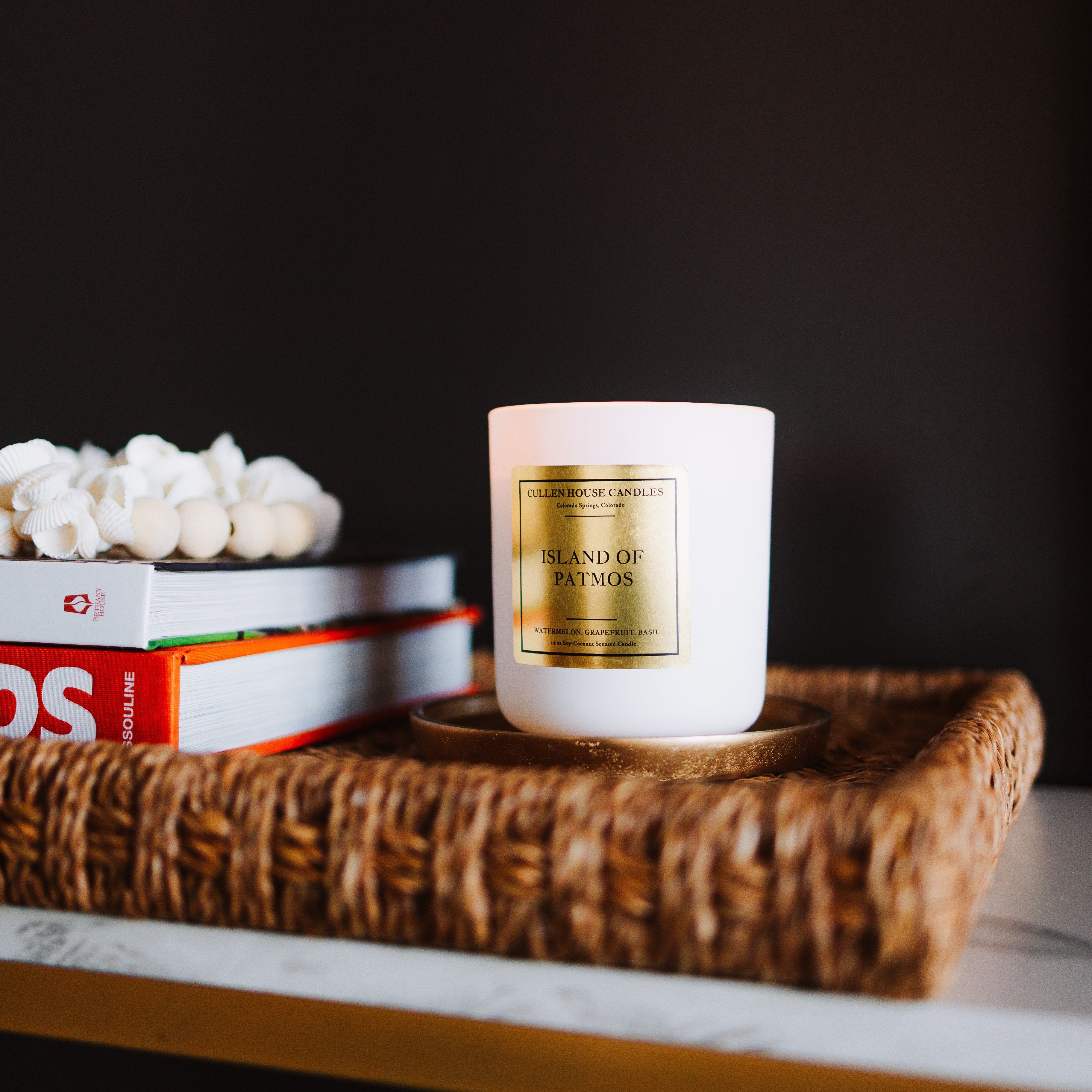 White candle with gold label on a woven tray with books against a dark background