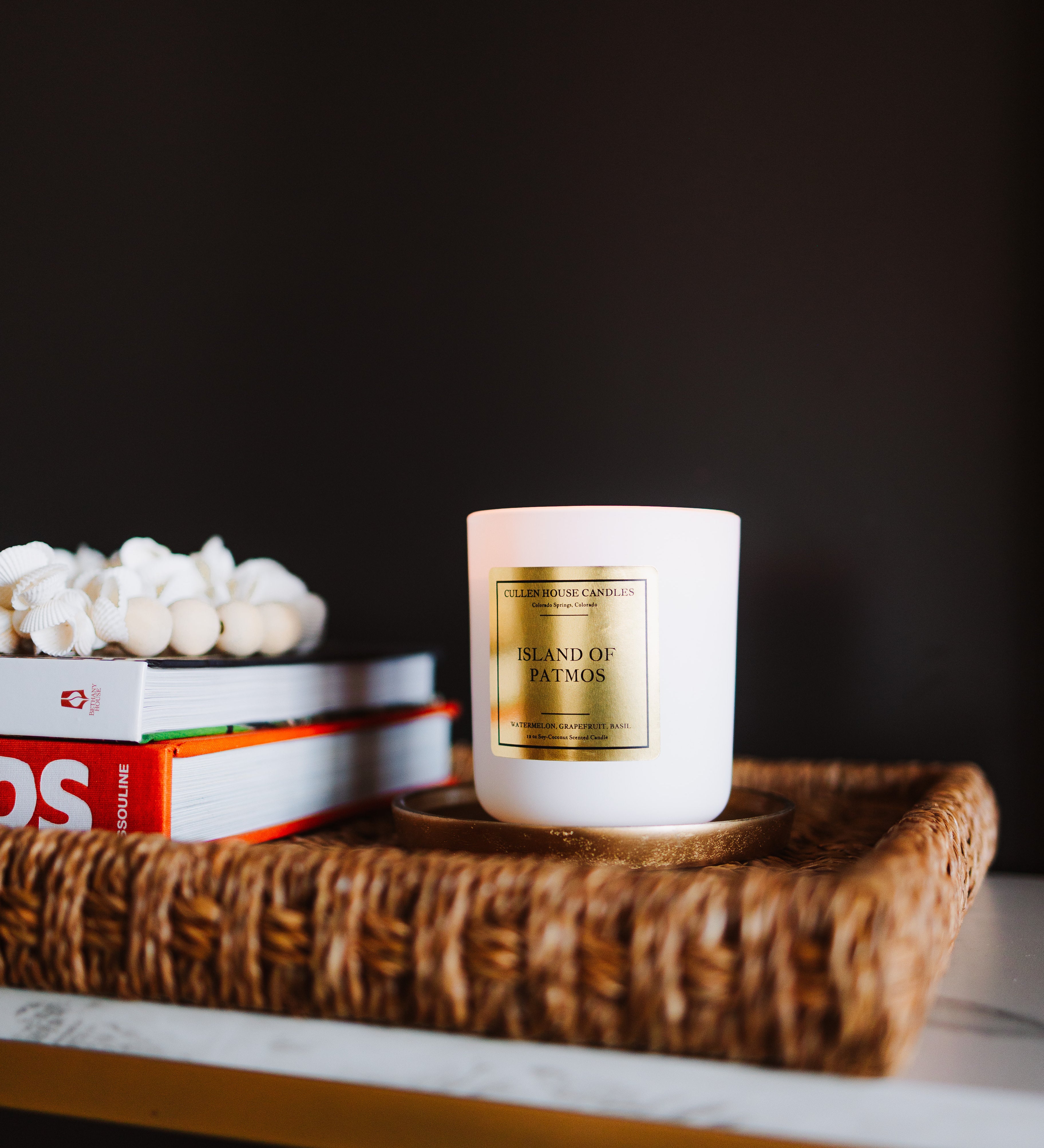 White candle with gold label on a woven tray with books against a dark background