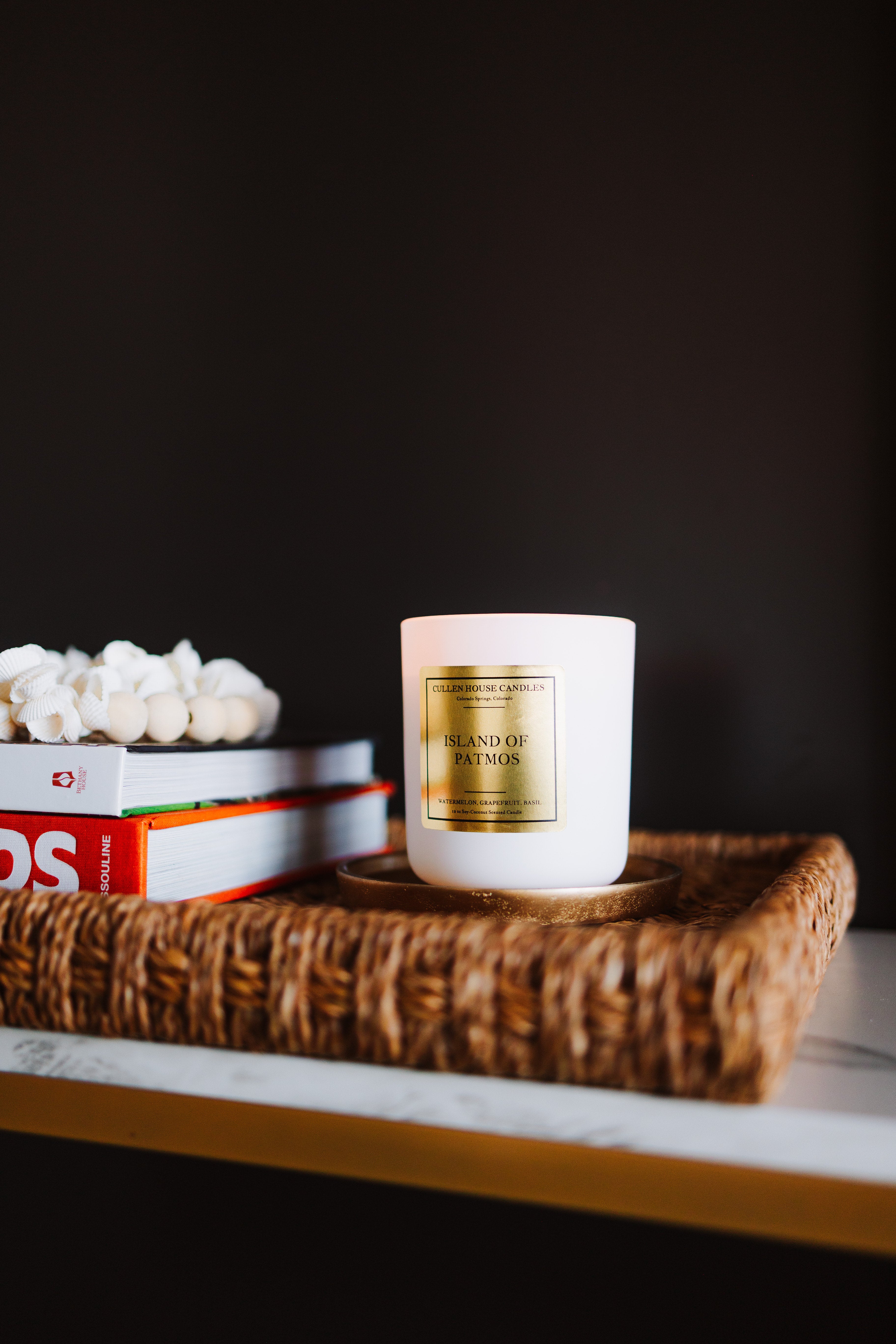 White candle with gold label on a woven tray with books against a dark background