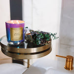 Candle with a purple and gold label on a small round table next to a sink.