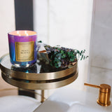 Candle with a purple and gold label on a small round table next to a sink.