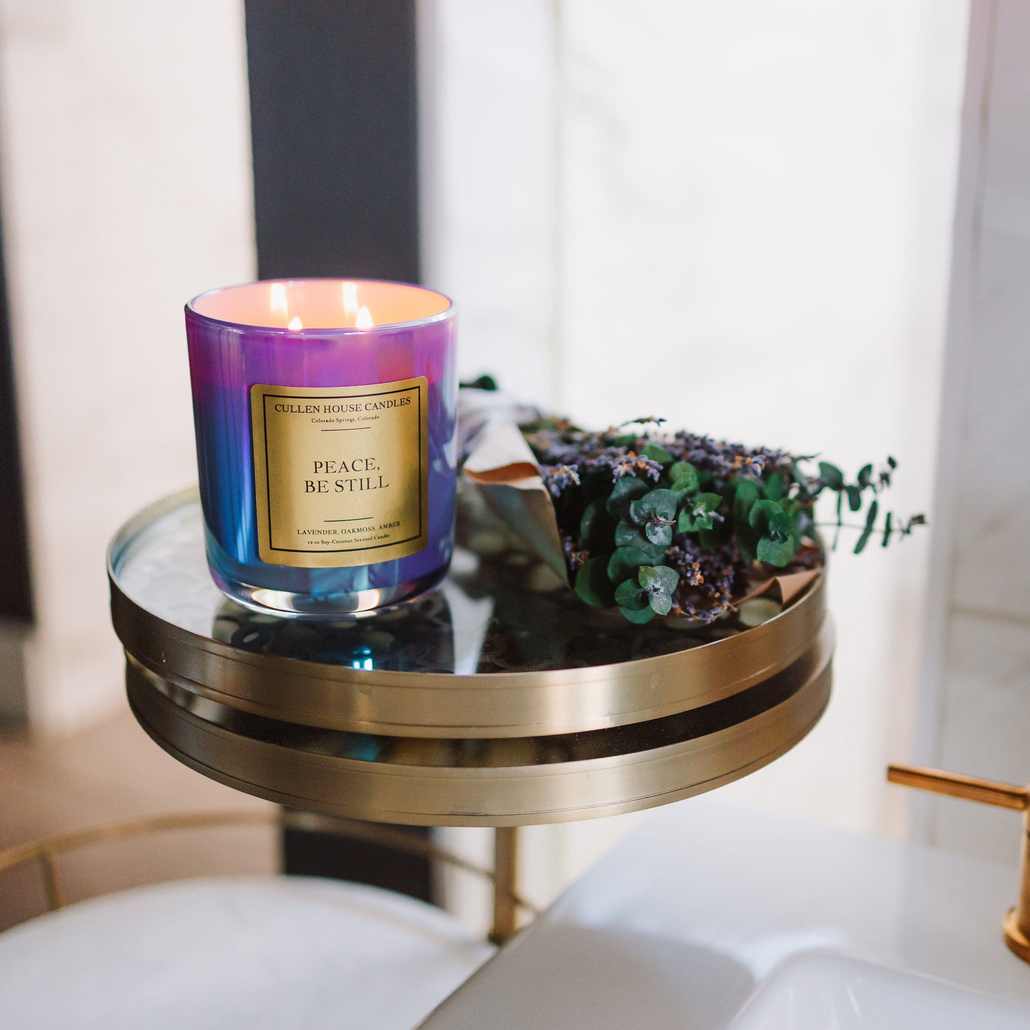 Candle with a purple and gold label on a small round table next to a sink.