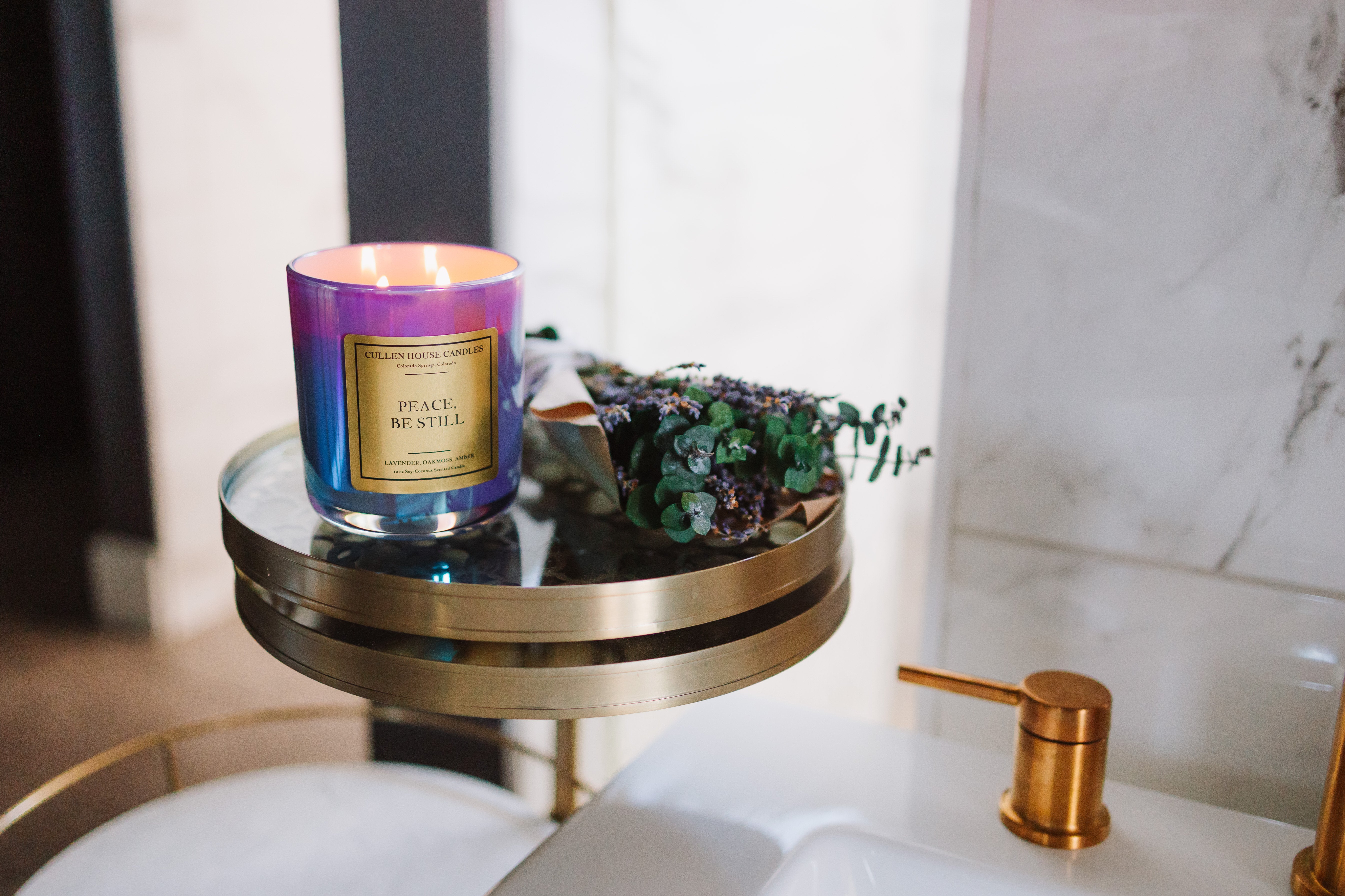Candle with a purple and gold label on a small round table next to a sink.