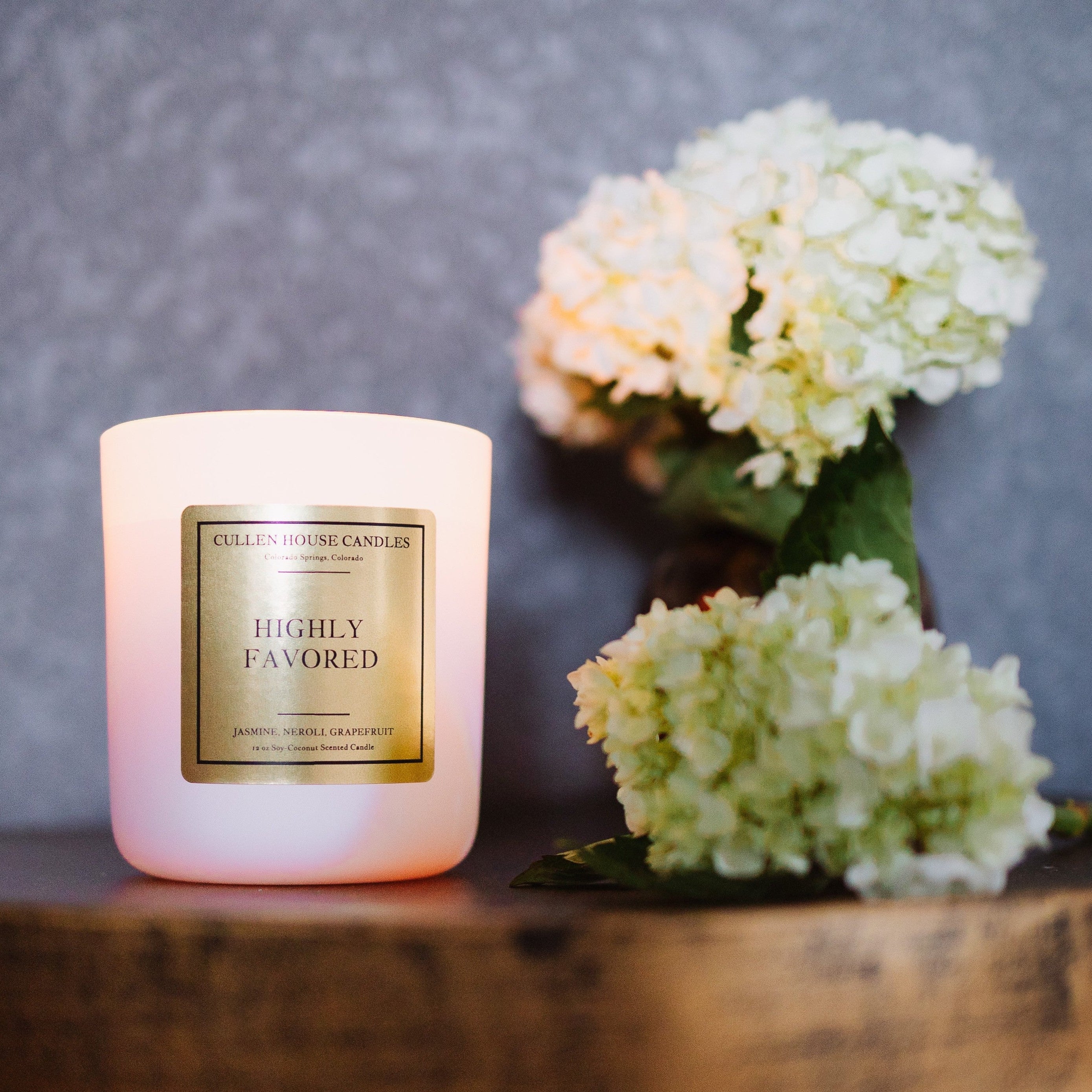 Pink candle labeled 'Highly Favored' with white flowers on a wooden surface