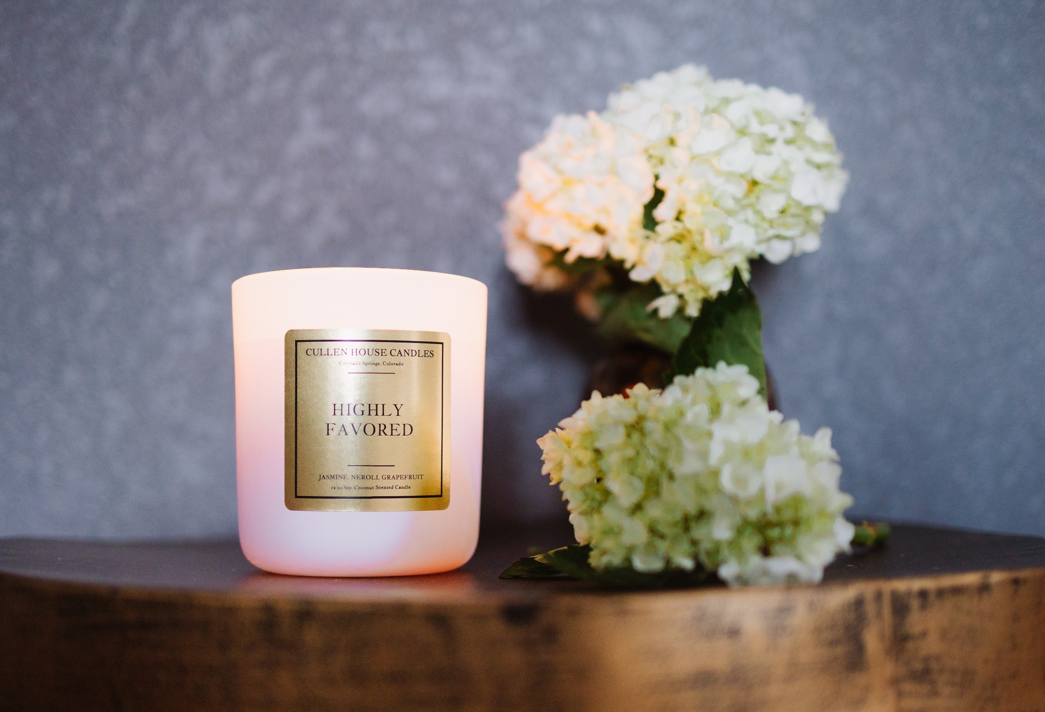 Pink candle labeled 'Highly Favored' with white flowers on a wooden surface