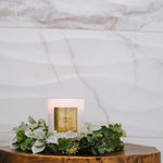 Candle labeled 'Lamb of God' on a wooden surface with greenery, against a marble background