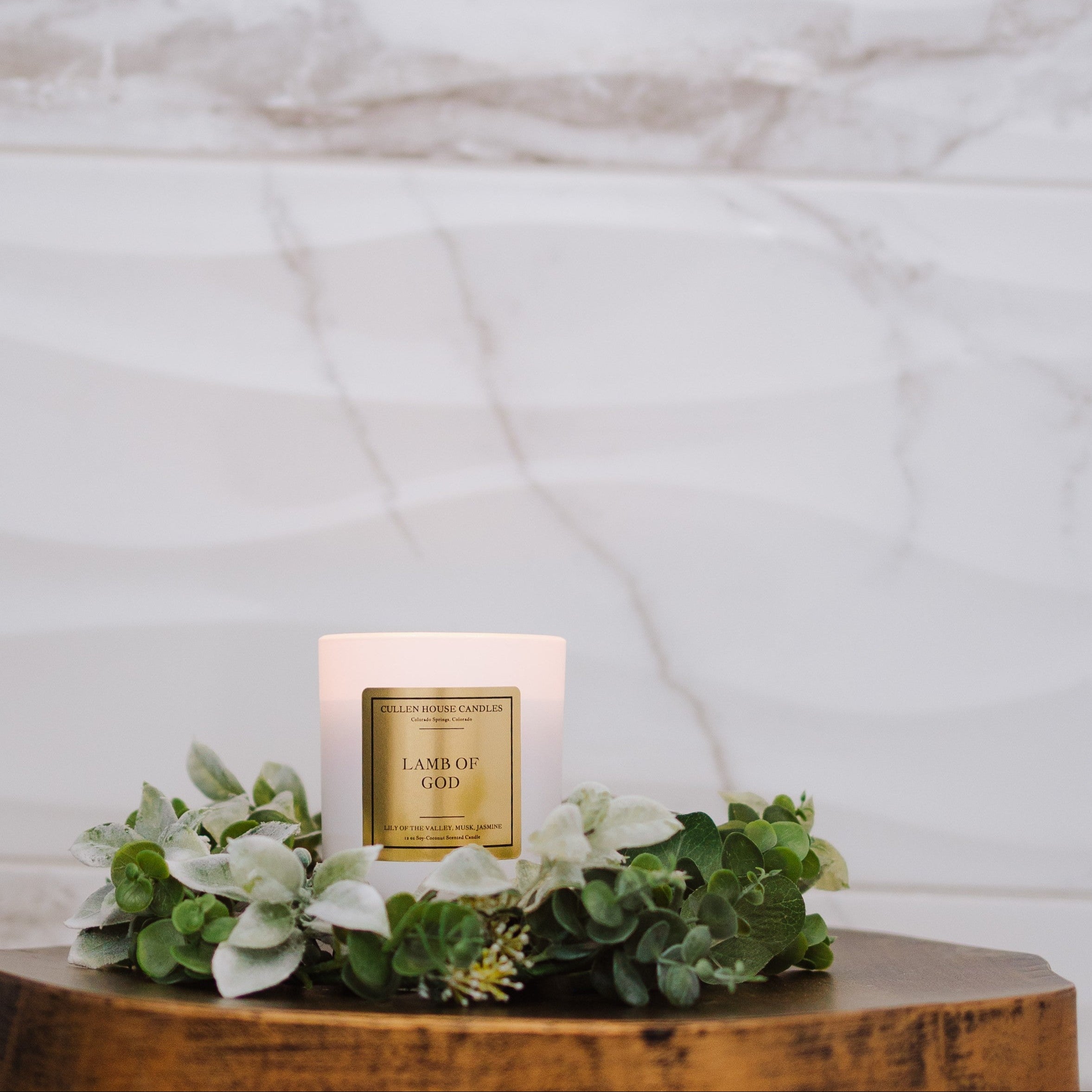 Candle labeled 'Lamb of God' on a wooden surface with greenery, against a marble background