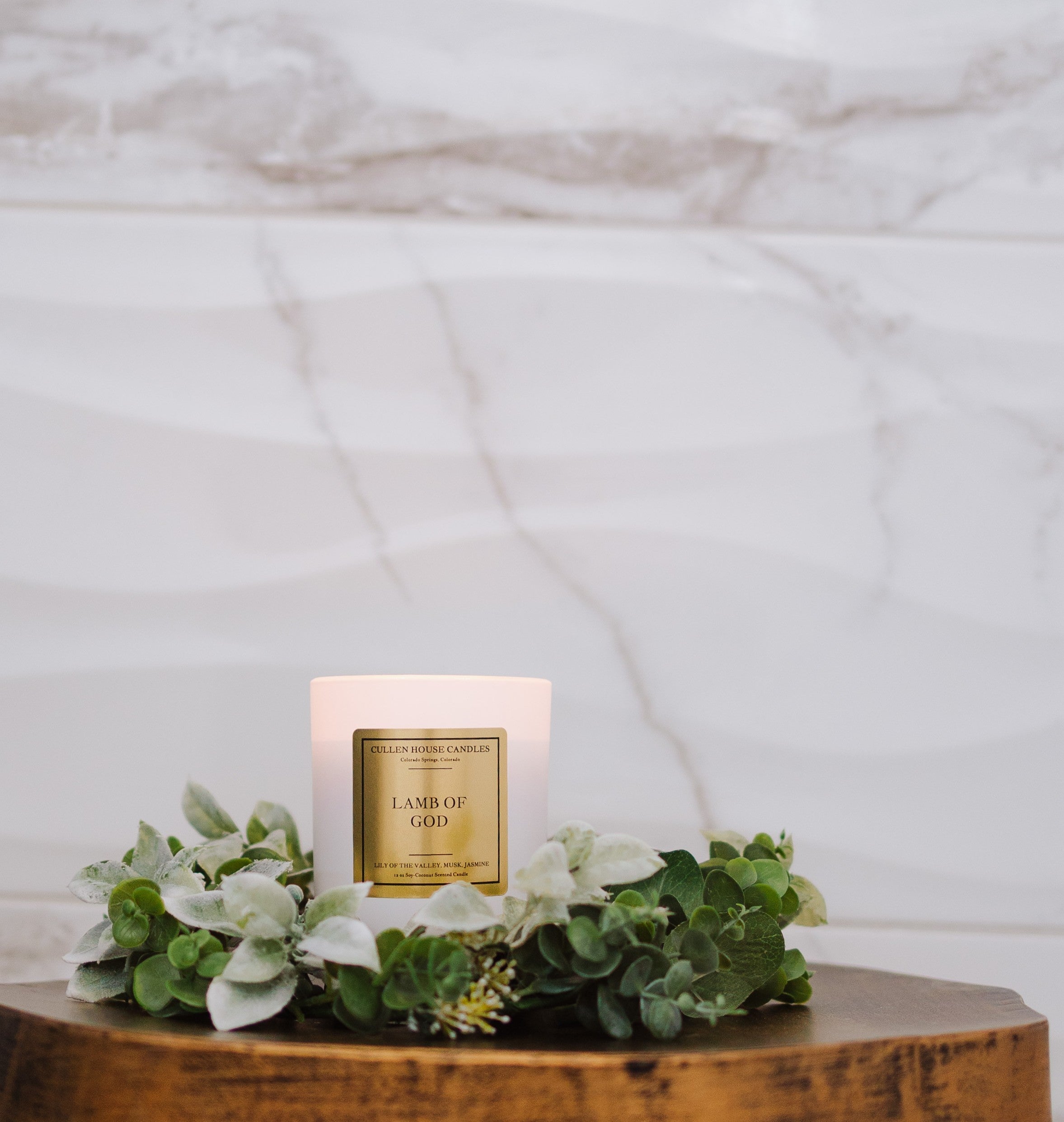 Candle labeled 'Lamb of God' on a wooden surface with greenery, against a marble background